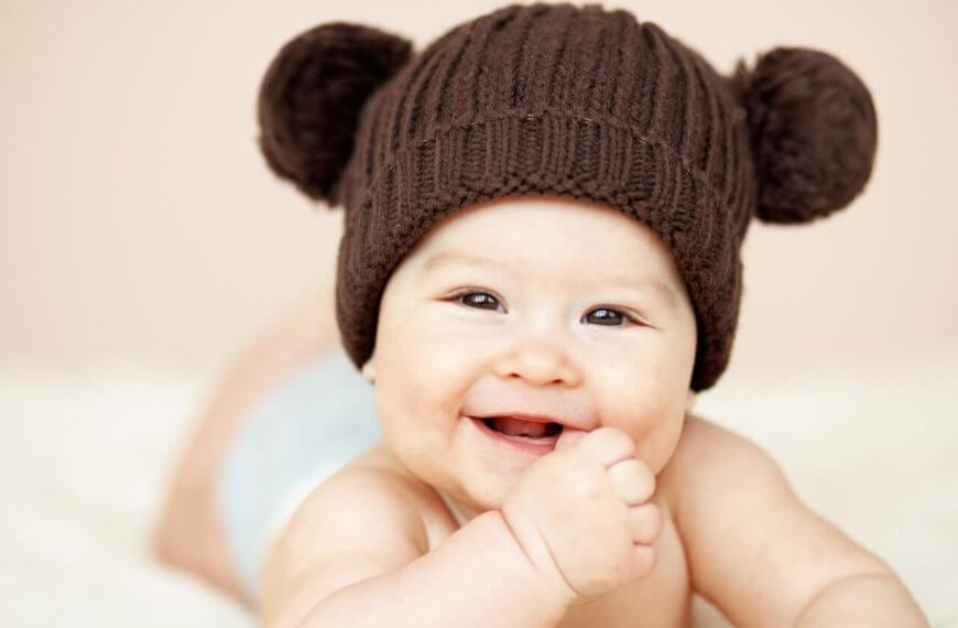 A smiling baby lies on their stomach, wearing a brown knitted hat with two pom-poms resembling bear ears. The baby’s hand is near their mouth, and they have a light-colored diaper on. The background is soft beige, adding to the warm and cozy feel of the image—perfect for showcasing baby essentials from the best baby stores.