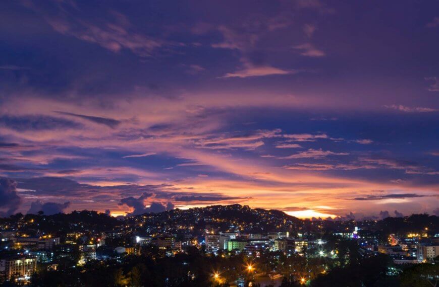 A panoramic view of a city at dusk with a vibrant, multicolored sky. The horizon showcases purple, pink, and orange hues as the sun sets. City lights flicker below, illuminating buildings and streets. Silhouetted clouds and hills add depth to the scene. The atmosphere is calm and picturesque—the perfect backdrop for Baguio Vacation Rentals or Budget Stays in Baguio Homes.