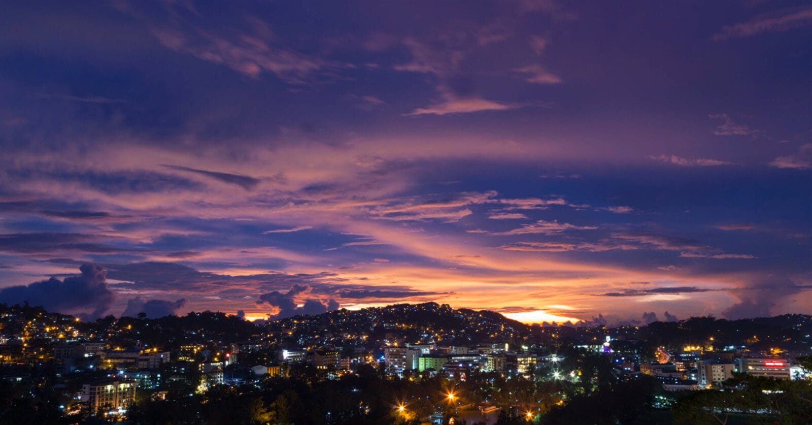 A panoramic view of a city at dusk with a vibrant, multicolored sky. The horizon showcases purple, pink, and orange hues as the sun sets. City lights flicker below, illuminating buildings and streets. Silhouetted clouds and hills add depth to the scene. The atmosphere is calm and picturesque—the perfect backdrop for Baguio Vacation Rentals or Budget Stays in Baguio Homes.