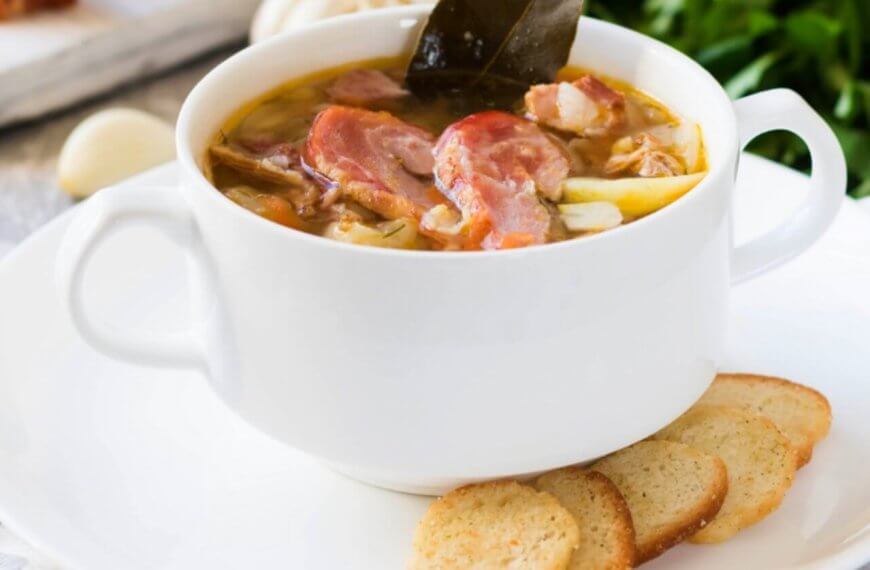 A white bowl filled with a hearty soup reminiscent of Tagaytay's famous Bulalo, containing chunks of meat, bone and vegetables, garnished with a leaf, is set on a white plate with three slices of toasted bread on the side. The background features ingredients like garlic and fresh greens, hinting at a rustic, homemade meal often celebrated in restaurants.