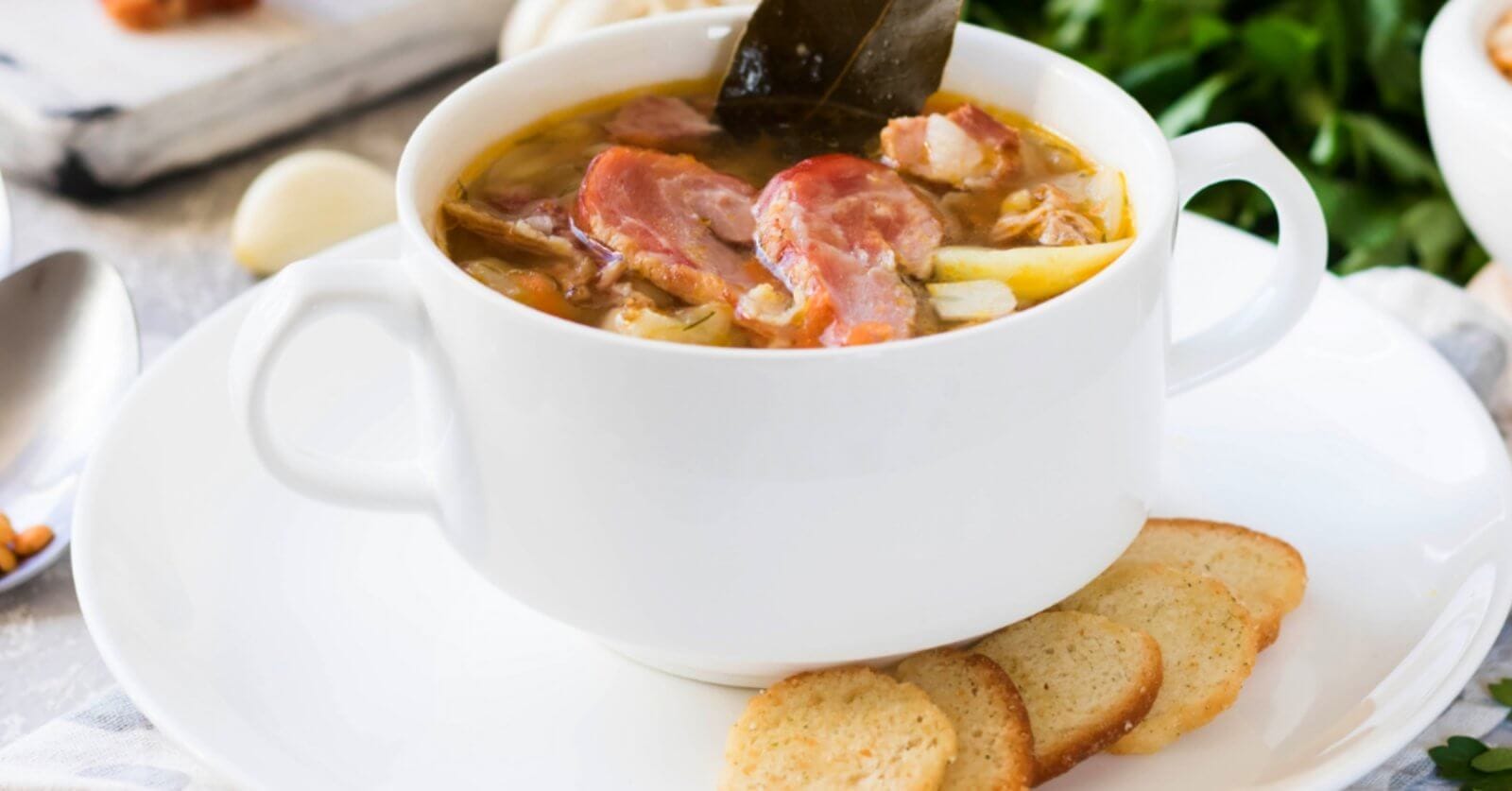 A white bowl filled with a hearty soup reminiscent of Tagaytay's famous Bulalo, containing chunks of meat, bone and vegetables, garnished with a leaf, is set on a white plate with three slices of toasted bread on the side. The background features ingredients like garlic and fresh greens, hinting at a rustic, homemade meal often celebrated in restaurants.