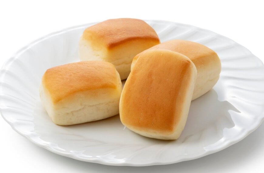 A white plate holds four golden-brown dinner rolls, each with a soft, fluffy texture. The rolls are slightly rectangular with rounded edges, and their tops are a light golden color that gradually lightens toward the bottom. Reminiscent of the best hot pandesal from Metro Manila's must-try bakeries, this plate features subtle decorative ridges around the rim.