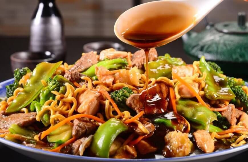 A close-up of a plate of stir-fried noodles loaded with vegetables like broccoli, bell peppers, and carrots, alongside sliced meat and shrimp. A hand is pouring a dark, glossy sauce from a spoon over the dish. In the background, two small bottles and a teapot are visible on a blurred dark surface—a scene reminiscent of the best Binondo restaurants.