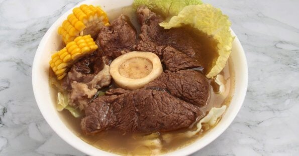 A bowl of hearty Bulalo soup featuring large chunks of tender beef, a central beef bone with marrow, corn on the cob pieces, and napa cabbage leaves in a clear broth. This Filipino favorite, popular in Metro Manila hotspots, is served in a white bowl set on a marble surface.