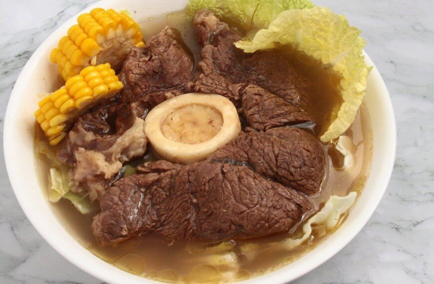A bowl of hearty Bulalo soup featuring large chunks of tender beef, a central beef bone with marrow, corn on the cob pieces, and napa cabbage leaves in a clear broth. This Filipino favorite, popular in Metro Manila hotspots, is served in a white bowl set on a marble surface.