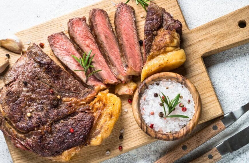 Grilled steak sliced into pieces on a wooden cutting board with a handle. Black and red pepper sprinkled on the steak. A wooden bowl filled with coarse salt and peppercorns sits next to the steak. Garnished with rosemary sprigs, garlic cloves, and accompanied by a fork and knife on the side—an experience at one of Eastwood City's must-visit restaurants.