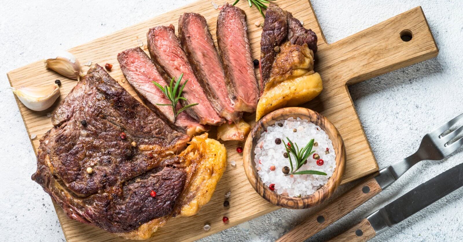 Grilled steak sliced into pieces on a wooden cutting board with a handle. Black and red pepper sprinkled on the steak. A wooden bowl filled with coarse salt and peppercorns sits next to the steak. Garnished with rosemary sprigs, garlic cloves, and accompanied by a fork and knife on the side—an experience at one of Eastwood City's must-visit restaurants.