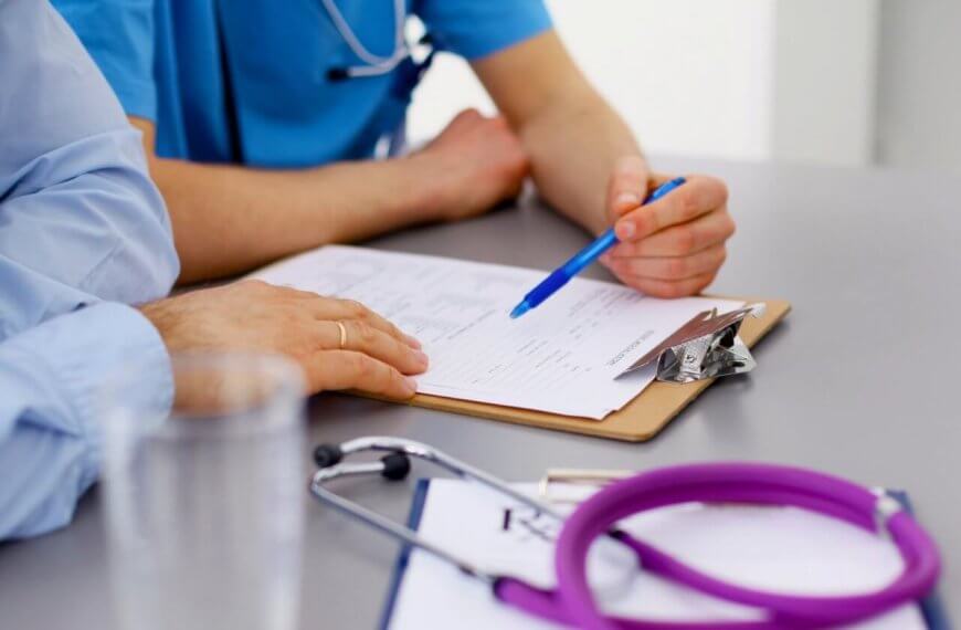 A close-up image of two family medicine doctors in Metro Manila reviewing a document on a clipboard. One person is holding a blue pen and pointing at the paper. The table has a stethoscope and additional papers. Both individuals are wearing medical scrubs, and there's a glass of water in the foreground.