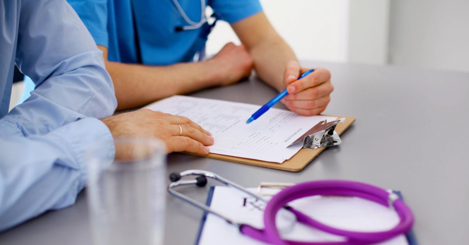A close-up image of two family medicine doctors in Metro Manila reviewing a document on a clipboard. One person is holding a blue pen and pointing at the paper. The table has a stethoscope and additional papers. Both individuals are wearing medical scrubs, and there's a glass of water in the foreground.