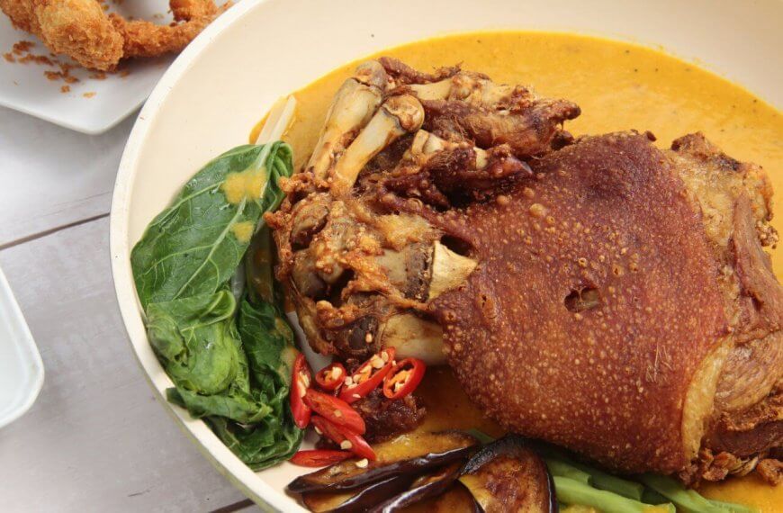 A must-try dish featuring crispy, deep-fried pork knuckle sits in a yellow curry sauce, accompanied by vegetables like leafy greens, eggplant slices, and red chili peppers. In the background, breaded shrimp and a small dish of dipping sauce are partially visible on white plates—an experience you can't miss at Filipino restaurants.