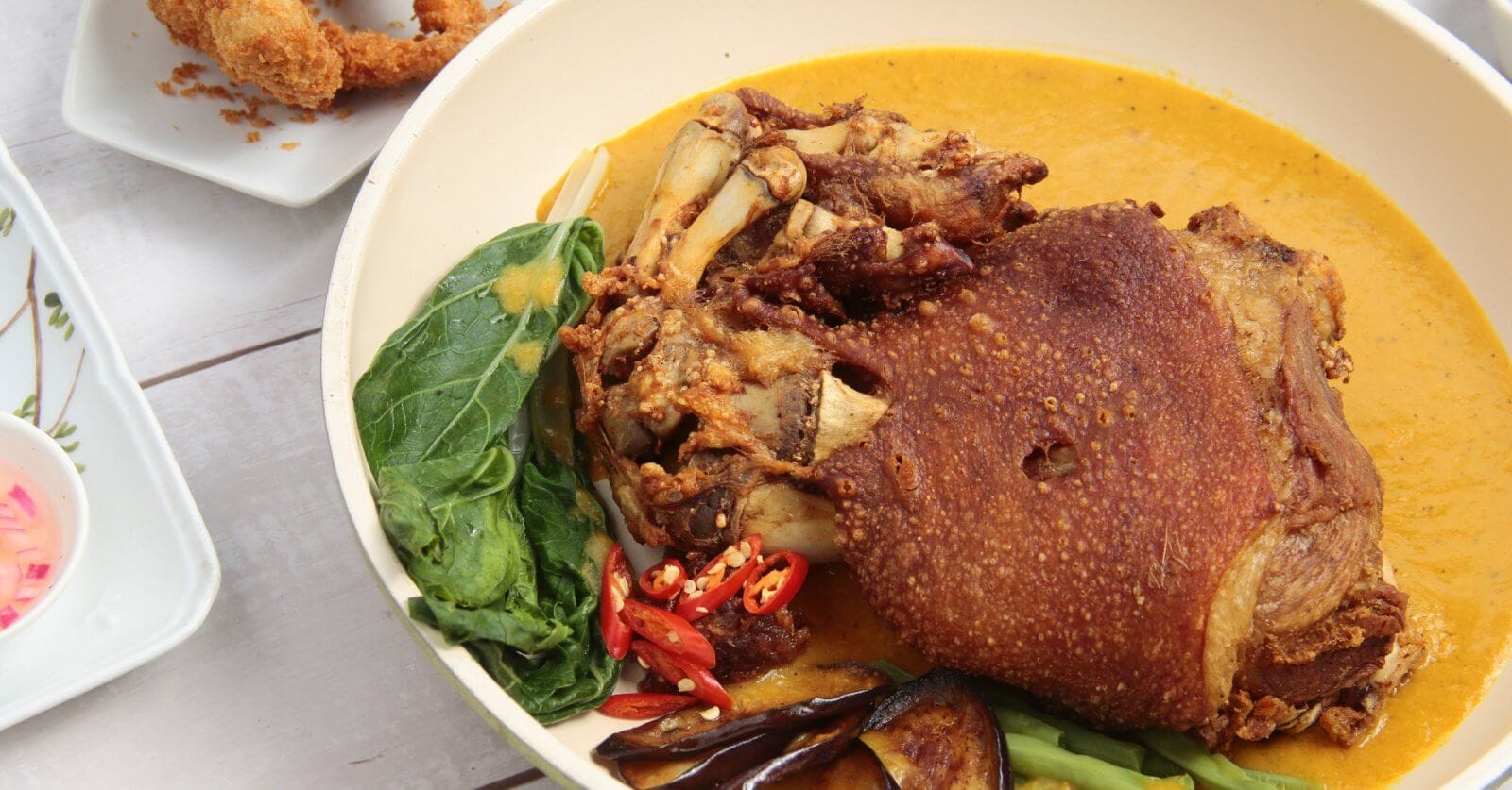 A must-try dish featuring crispy, deep-fried pork knuckle sits in a yellow curry sauce, accompanied by vegetables like leafy greens, eggplant slices, and red chili peppers. In the background, breaded shrimp and a small dish of dipping sauce are partially visible on white plates—an experience you can't miss at Filipino restaurants.