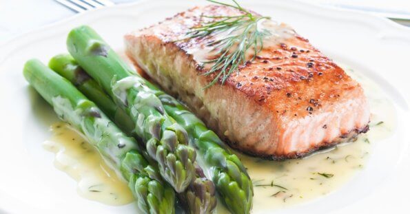 A plate of food featuring a grilled salmon fillet garnished with fresh dill, accompanied by several spears of steamed asparagus. The dish is drizzled with a creamy dill sauce, showcasing the best Greenhills dining experience. The plate sits on a white tablecloth with a fork partially visible in the background.
