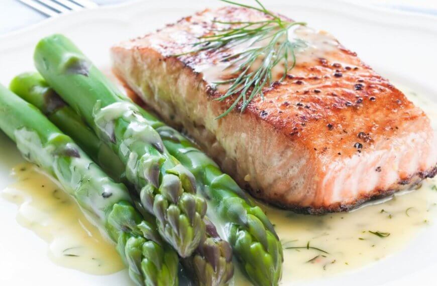 A plate of food featuring a grilled salmon fillet garnished with fresh dill, accompanied by several spears of steamed asparagus. The dish is drizzled with a creamy dill sauce, showcasing the best Greenhills dining experience. The plate sits on a white tablecloth with a fork partially visible in the background.