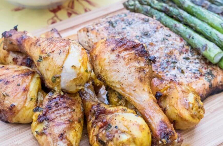 Grilled chicken drumsticks, seasoned and charred, are arranged in the foreground on a wooden board, alongside a piece of herb-topped inihaw fish. To the side, there's a row of grilled asparagus spears, adding a vibrant green contrast to this delightful dish reminiscent of popular hotspots in Metro Manila.