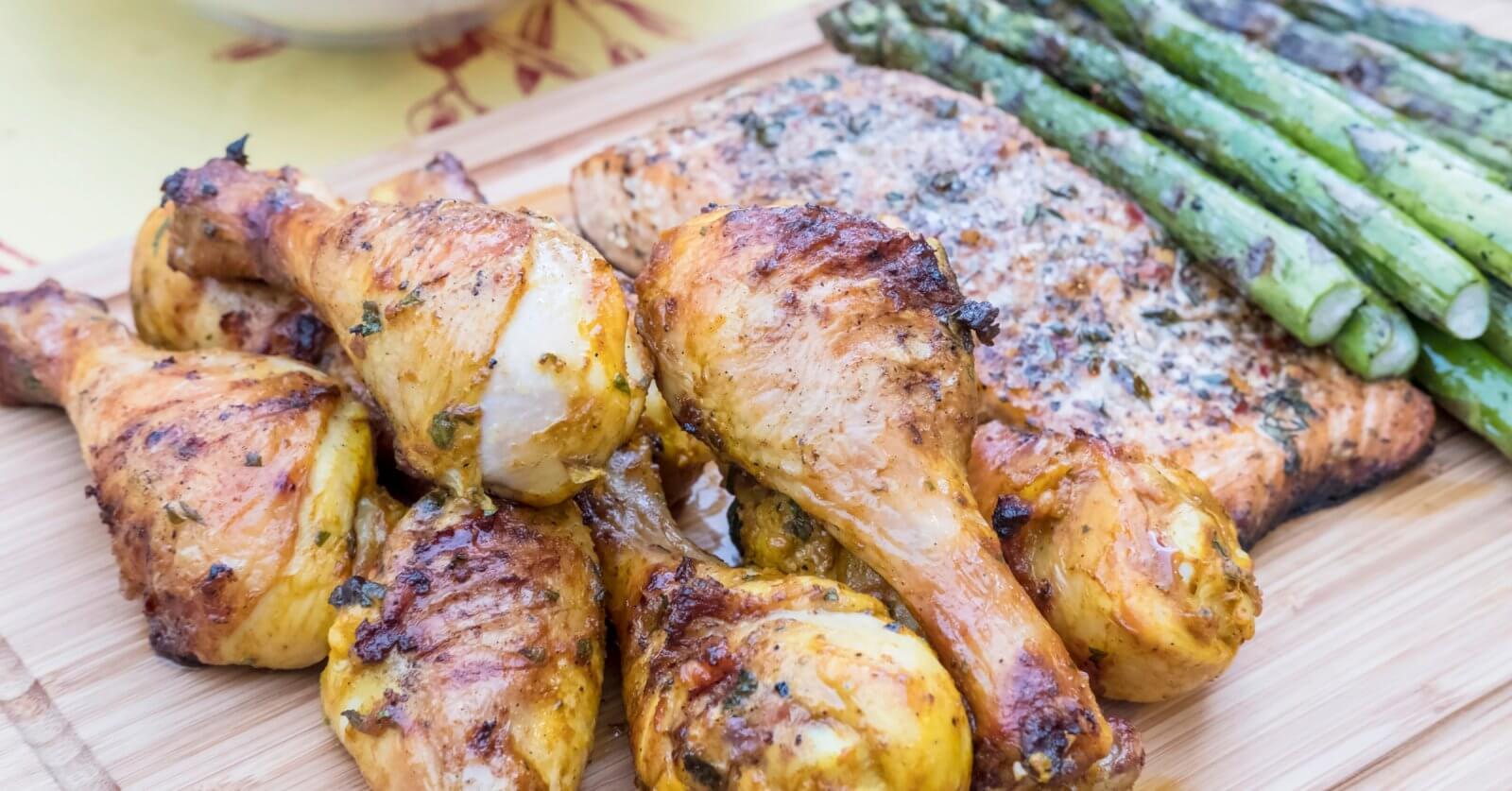 Grilled chicken drumsticks, seasoned and charred, are arranged in the foreground on a wooden board, alongside a piece of herb-topped inihaw fish. To the side, there's a row of grilled asparagus spears, adding a vibrant green contrast to this delightful dish reminiscent of popular hotspots in Metro Manila.