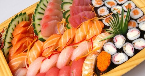 A large wooden platter filled with an assortment of sushi and sashimi, reminiscent of offerings found in Japanese grocery stores across Metro Manila. The platter includes various kinds of fish like salmon, tuna, and white fish, garnished with thin vegetable slices and shredded carrots. There are also sushi rolls, some topped with orange roe, and garnished with a clump of green grass.