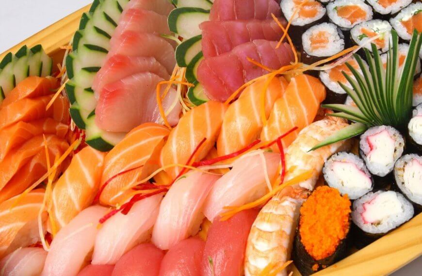 A large wooden platter filled with an assortment of sushi and sashimi, reminiscent of offerings found in Japanese grocery stores across Metro Manila. The platter includes various kinds of fish like salmon, tuna, and white fish, garnished with thin vegetable slices and shredded carrots. There are also sushi rolls, some topped with orange roe, and garnished with a clump of green grass.