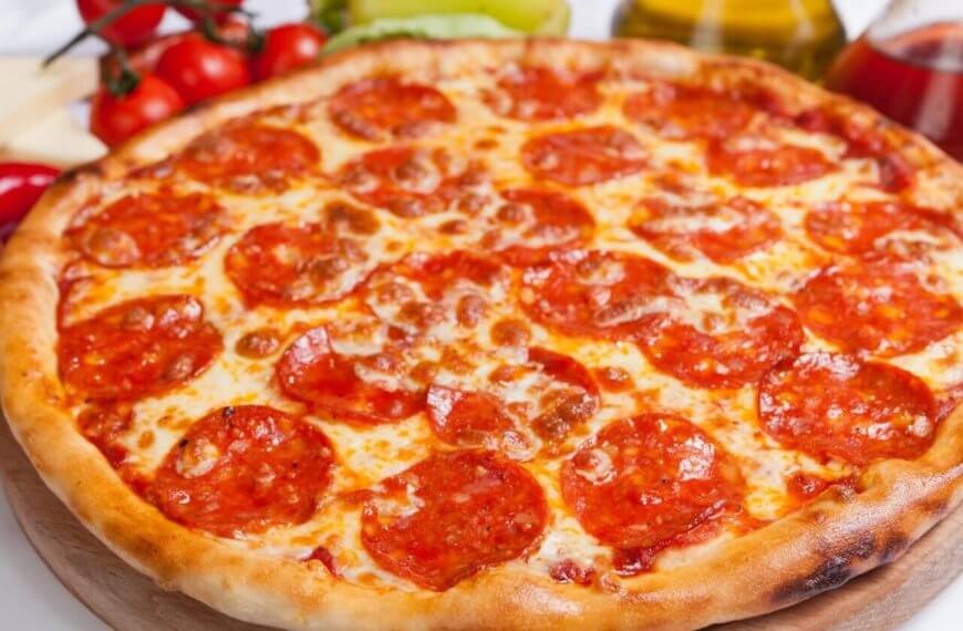 A freshly baked pepperoni pizza with a golden-brown crust is displayed on a wooden board. The pizza, reminiscent of what you might find at top restaurants, is topped with melted cheese and an uneven spread of pepperoni slices. Ingredients like tomatoes, peppers, cheese, and oil bottles are blurred in the background.