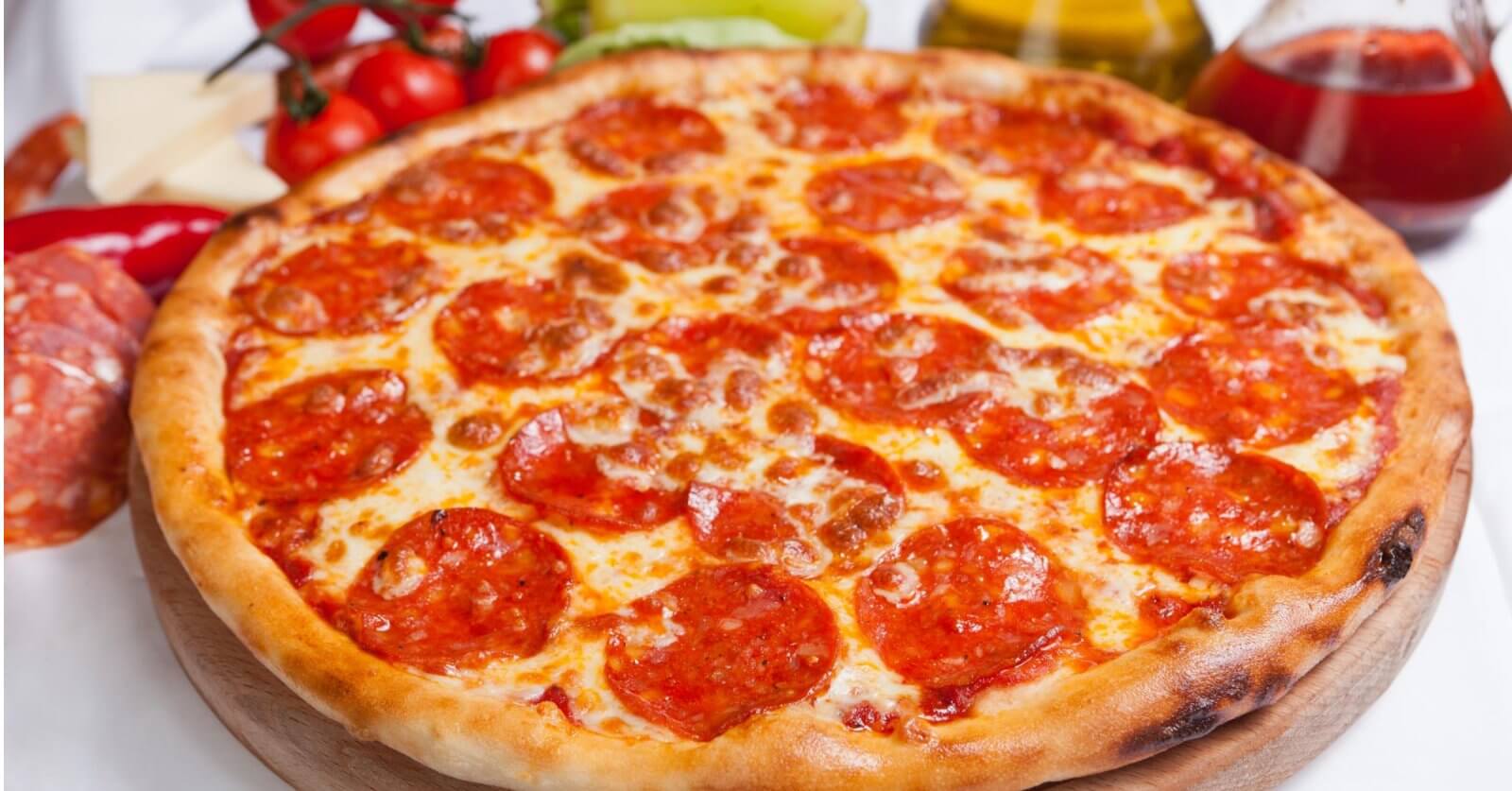 A freshly baked pepperoni pizza with a golden-brown crust is displayed on a wooden board. The pizza, reminiscent of what you might find at top restaurants, is topped with melted cheese and an uneven spread of pepperoni slices. Ingredients like tomatoes, peppers, cheese, and oil bottles are blurred in the background.