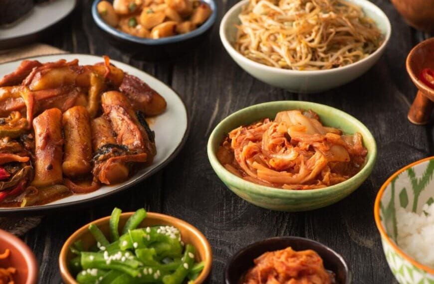 A variety of Korean dishes are displayed on a dark wooden table. The spread, which showcases the best of Korean cuisine, includes a plate of stir-fried meat and vegetables, a bowl of sauteed bean sprouts, kimchi from one of Metro Manila’s top Korean groceries, sliced red chili peppers, green beans with sesame seeds, white rice, and small dishes of other sides.