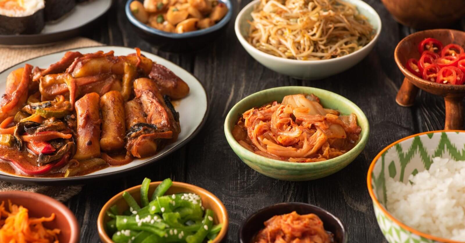 A variety of Korean dishes are displayed on a dark wooden table. The spread, which showcases the best of Korean cuisine, includes a plate of stir-fried meat and vegetables, a bowl of sauteed bean sprouts, kimchi from one of Metro Manila’s top Korean groceries, sliced red chili peppers, green beans with sesame seeds, white rice, and small dishes of other sides.