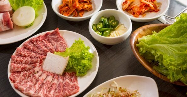 An array of Korean BBQ ingredients on a wooden table, featuring a plate of marbled beef slices with a square of tofu and lettuce, a platter of fresh lettuce leaves, and smaller bowls containing sliced onions, kimchi, green chili peppers, garlic cloves, and a mixed vegetable side dish—all part of the ultimate dining experience at top Korean restaurants in Metro Manila.