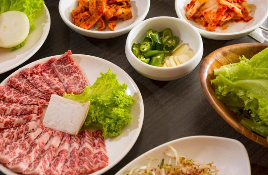 An array of Korean BBQ ingredients on a wooden table, featuring a plate of marbled beef slices with a square of tofu and lettuce, a platter of fresh lettuce leaves, and smaller bowls containing sliced onions, kimchi, green chili peppers, garlic cloves, and a mixed vegetable side dish—all part of the ultimate dining experience at top Korean restaurants in Metro Manila.
