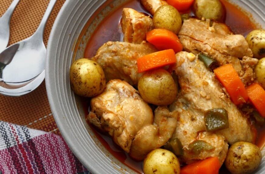 A savory dish of chicken stew is served in a grey bowl. The stew includes tender chicken pieces, whole baby potatoes, sliced carrots, and green bell pepper chunks, all immersed in a rich, red-colored broth. A fork and spoon are placed beside the bowl, resting on a colorful cloth napkin—an absolute must-try at Legaspi Village restaurants.