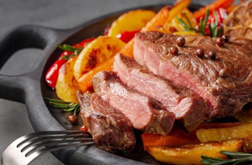 A juicy, medium-rare steak sliced into pieces sits on a black cast-iron skillet. The steak is garnished with whole peppercorns and fresh rosemary. Accompanying the steak are roasted potato wedges, red peppers, and a halved head of garlic. Nearby restaurants in Magallanes Village, Makati serve similar delights.