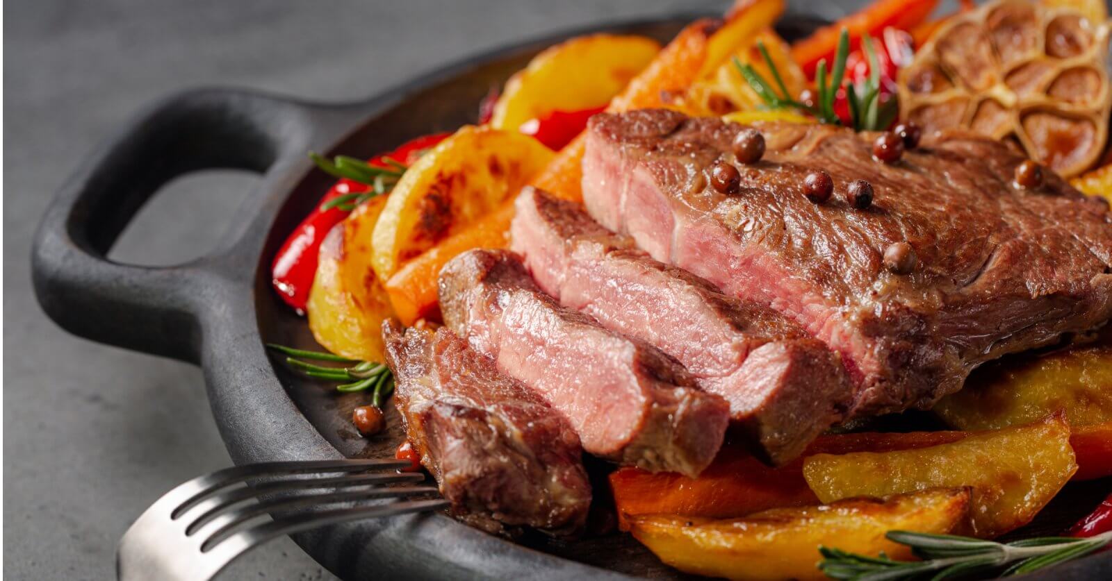 A juicy, medium-rare steak sliced into pieces sits on a black cast-iron skillet. The steak is garnished with whole peppercorns and fresh rosemary. Accompanying the steak are roasted potato wedges, red peppers, and a halved head of garlic. Nearby restaurants in Magallanes Village, Makati serve similar delights.