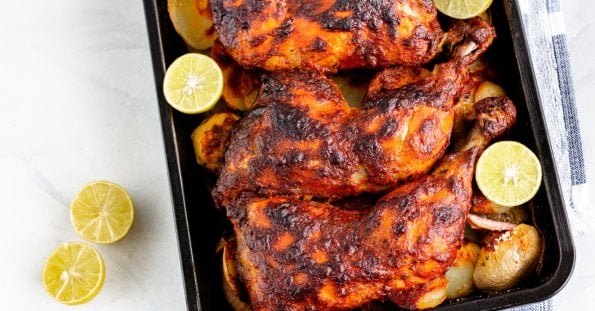 Three roasted chicken legs are placed on a baking tray, surrounded by roasted potato slices and garnished with halved limes. The chicken legs are marinated with a dark, crusty seasoning. Two halved limes rest on the white countertop next to the tray, reminiscent of dishes from restaurants along Marcos Highway.
