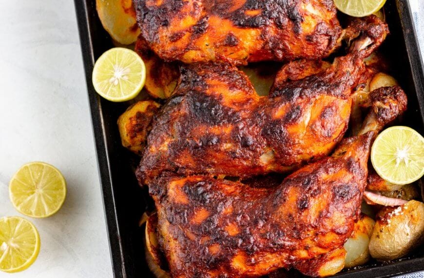 Three roasted chicken legs are placed on a baking tray, surrounded by roasted potato slices and garnished with halved limes. The chicken legs are marinated with a dark, crusty seasoning. Two halved limes rest on the white countertop next to the tray, reminiscent of dishes from restaurants along Marcos Highway.