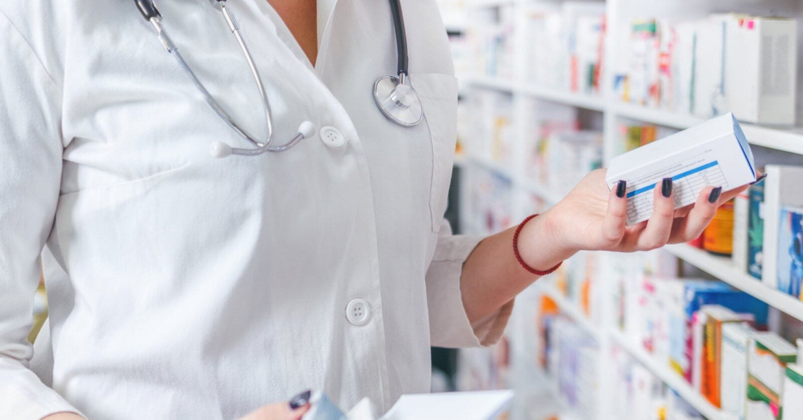 A pharmacist wearing a white coat and stethoscope is standing in a Mercury Branches pharmacy in Metro Manila. They are holding two boxes of medication while reading the label on one. Shelves filled with various medicine boxes are visible in the background.