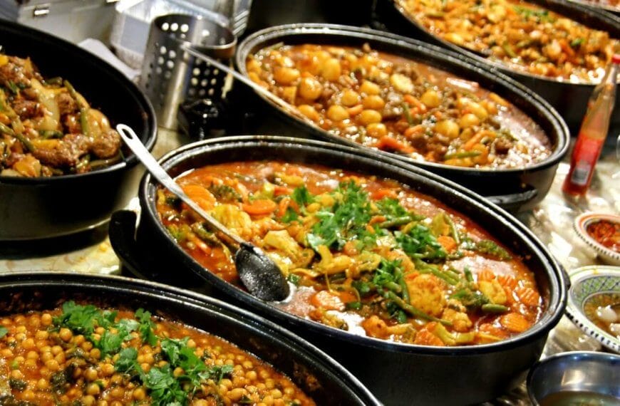 A hearty spread of various cooked dishes in large black pots is arranged on a table at the Eat-All-You-Can Buffet in SM Mall of Asia, featuring chickpea stew, meat and vegetable curry, and a tomato-based stew with herbs. Small bowls containing condiments and garnishes surround the main dishes, with serving spoons resting in the pots.