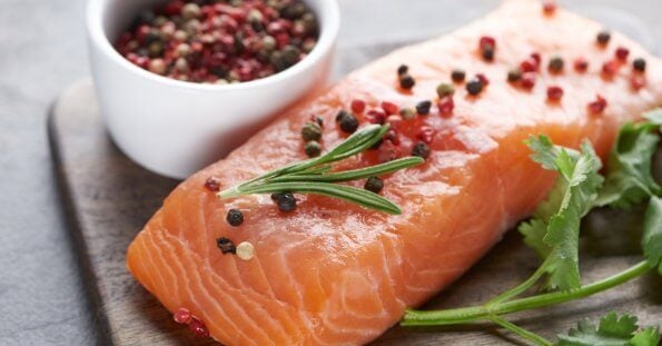 A raw salmon fillet is placed on a wooden surface, garnished with rosemary and an assortment of whole peppercorns. A small white bowl filled with mixed peppercorns is next to the salmon. Fresh cilantro sprigs lie beside the fillet, adding a touch of green to the presentation—a scene reminiscent of dishes at Molito Lifestyle Center's best restaurants.