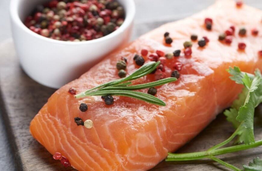 A raw salmon fillet is placed on a wooden surface, garnished with rosemary and an assortment of whole peppercorns. A small white bowl filled with mixed peppercorns is next to the salmon. Fresh cilantro sprigs lie beside the fillet, adding a touch of green to the presentation—a scene reminiscent of dishes at Molito Lifestyle Center's best restaurants.