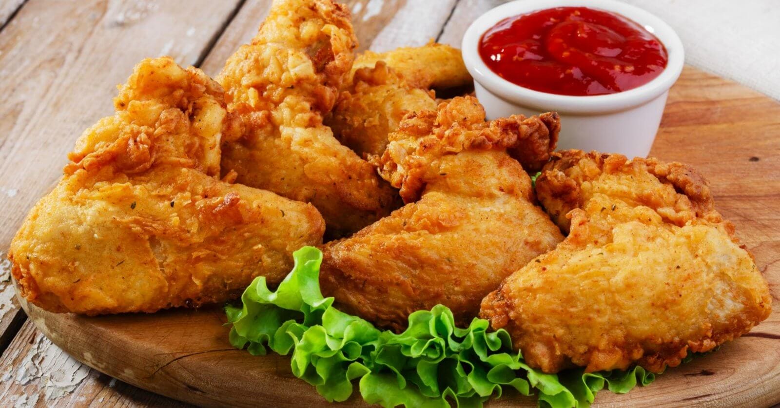 Close-up of crispy, golden-brown fried chicken pieces arranged on a wooden board, garnished with fresh green lettuce leaves at the bottom. A small white cup filled with red ketchup is placed behind the chicken. This must-try dish, found in Parañaque restaurants, sits against a rustic wooden background.
