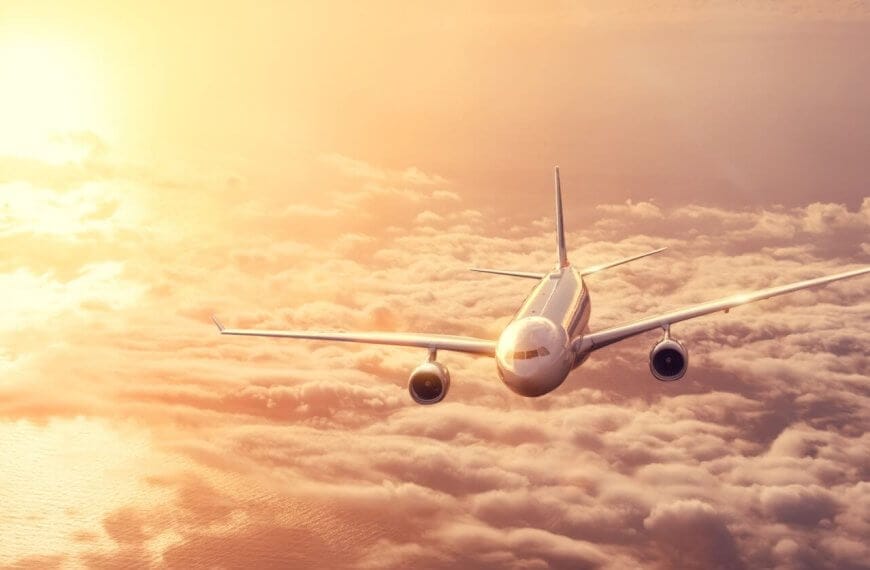 A commercial airplane from Philippine Airlines flies above a dense layer of clouds during sunset. The sky is filled with warm hues of orange and yellow, casting a golden glow on the plane and clouds. The airplane is heading toward the camera, with both wings and engines visible.