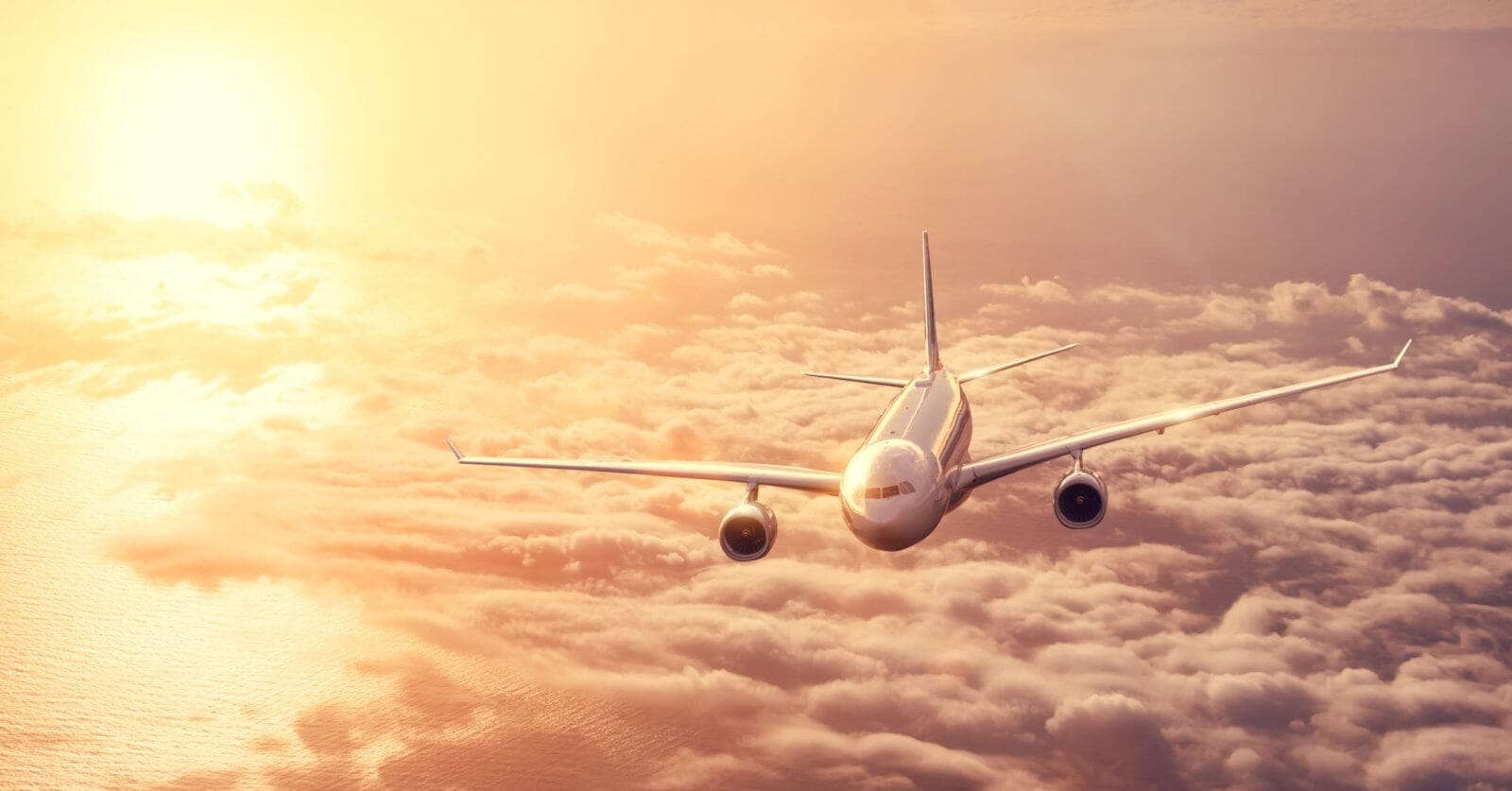 A commercial airplane from Philippine Airlines flies above a dense layer of clouds during sunset. The sky is filled with warm hues of orange and yellow, casting a golden glow on the plane and clouds. The airplane is heading toward the camera, with both wings and engines visible.