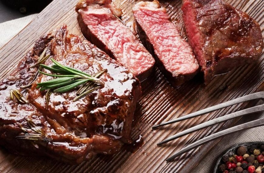A wooden cutting board with a juicy, medium-rare steak is garnished with fresh rosemary and sliced into three pieces. A vintage fork rests on the board, beside a small bowl of colorful peppercorns. The neutral-toned cloth backdrop lends a charming vibe, reminiscent of delightful dining spots in Salcedo Village.