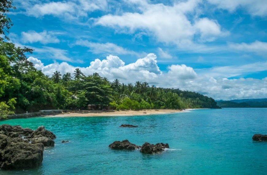 A serene tropical beach with clear turquoise waters, scattered rocks, and lush green vegetation along the shoreline. The sky is bright blue with scattered white clouds. Palm trees and dense forest extend to the edge of the sandy beach at one of the Best Samal Islands Resorts, creating a picturesque coastal landscape.