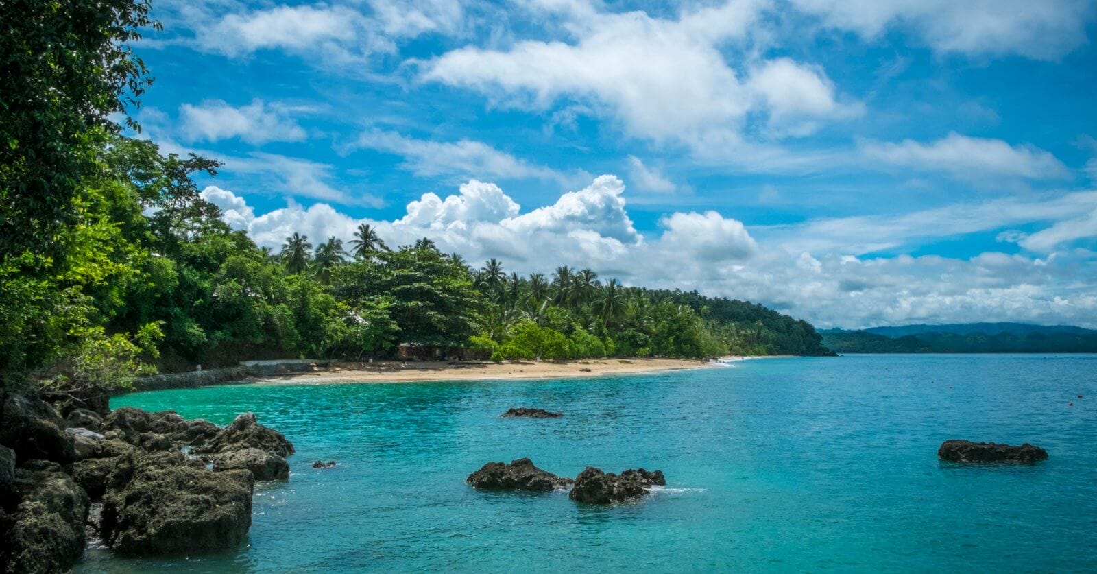 A serene tropical beach with clear turquoise waters, scattered rocks, and lush green vegetation along the shoreline. The sky is bright blue with scattered white clouds. Palm trees and dense forest extend to the edge of the sandy beach at one of the Best Samal Islands Resorts, creating a picturesque coastal landscape.