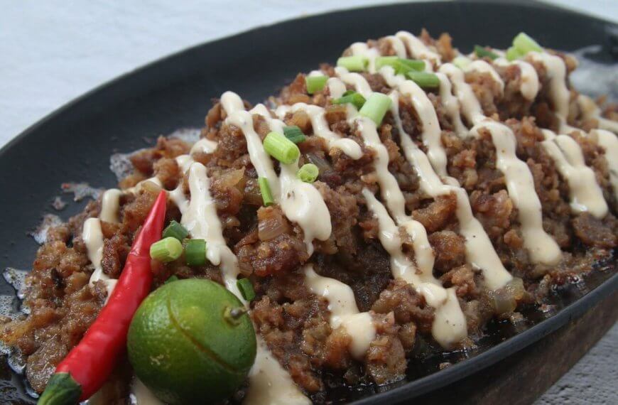 A sizzling platter of sisig is topped with creamy mayonnaise drizzles and garnished with chopped green onions. A whole red chili pepper and a halved calamansi adorn the side, enhancing the dish's vibrant presentation. This culinary delight, perfect to visit at San Fernando restaurants, is set against a neutral background.