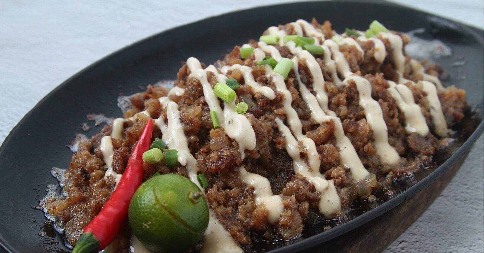 A sizzling platter of sisig is topped with creamy mayonnaise drizzles and garnished with chopped green onions. A whole red chili pepper and a halved calamansi adorn the side, enhancing the dish's vibrant presentation. This culinary delight, perfect to visit at San Fernando restaurants, is set against a neutral background.