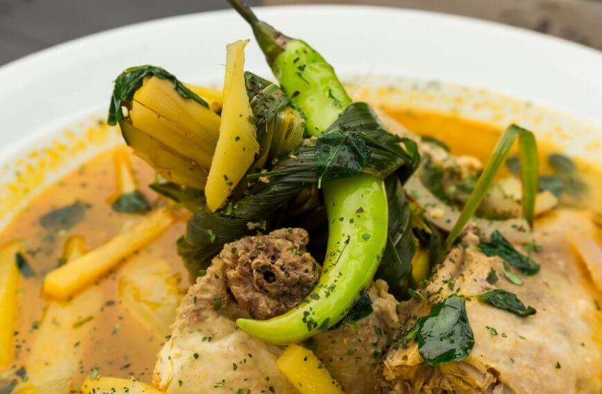 A close-up of a bowl of soup features tender meat pieces, green chili peppers, leafy greens, and yellow vegetable slices in a yellowish broth. The soup is garnished with herbs and served in a white bowl. The richly seasoned broth delivers an aromatic experience you'd expect from one of SM Megamall's best restaurants.