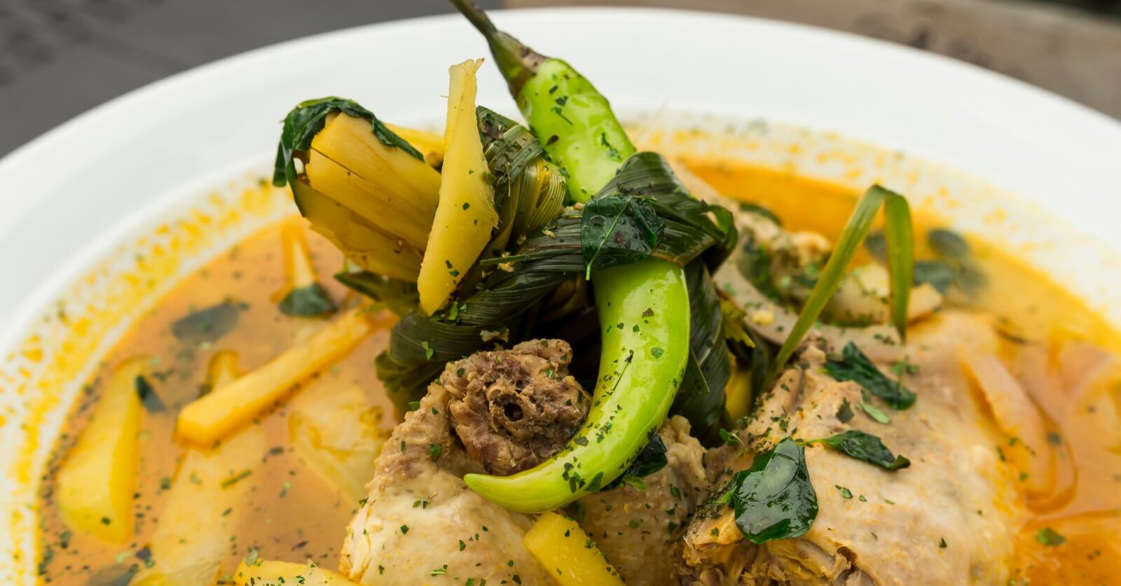 A close-up of a bowl of soup features tender meat pieces, green chili peppers, leafy greens, and yellow vegetable slices in a yellowish broth. The soup is garnished with herbs and served in a white bowl. The richly seasoned broth delivers an aromatic experience you'd expect from one of SM Megamall's best restaurants.