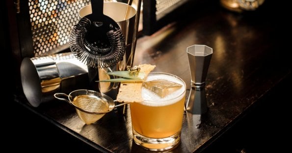 A well-lit bar setup in a hidden gem of Manila features a cocktail garnished with a pineapple leaf and dried pineapple slice on a wooden surface. Nearby are a strainer, shaker, jigger, and fine mesh sieve, evoking the professional mixology found in speakeasy bars.