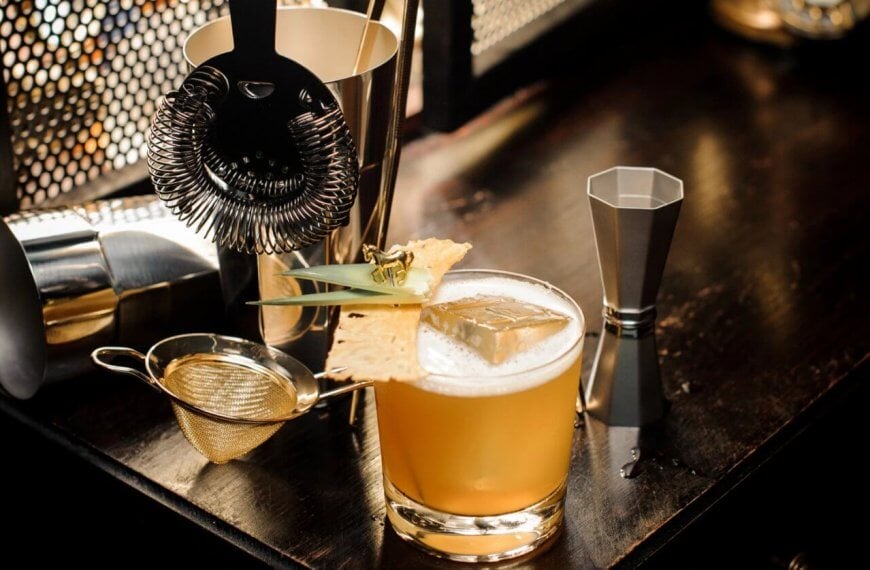 A well-lit bar setup in a hidden gem of Manila features a cocktail garnished with a pineapple leaf and dried pineapple slice on a wooden surface. Nearby are a strainer, shaker, jigger, and fine mesh sieve, evoking the professional mixology found in speakeasy bars.