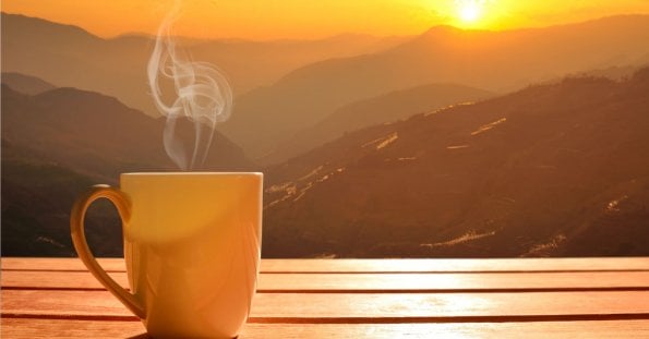 A steaming mug sits on a wooden surface, with a breathtaking sunrise over the mountain ranges of Tanay, Rizal in the background. The sky displays hues of orange and yellow, casting a warm glow over the landscape. Mist gently rises from the valleys, adding a serene and tranquil atmosphere to one of the best coffee spots you'll ever find.