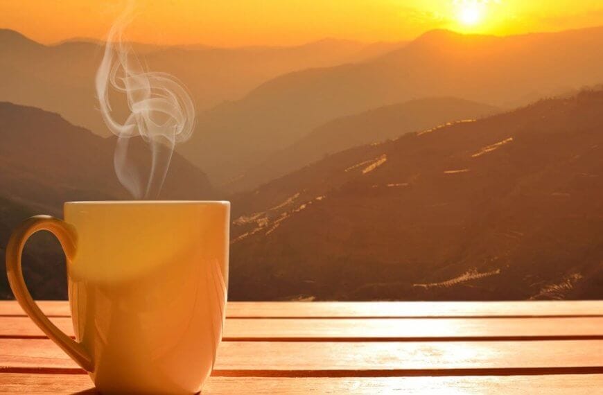 A steaming mug sits on a wooden surface, with a breathtaking sunrise over the mountain ranges of Tanay, Rizal in the background. The sky displays hues of orange and yellow, casting a warm glow over the landscape. Mist gently rises from the valleys, adding a serene and tranquil atmosphere to one of the best coffee spots you'll ever find.